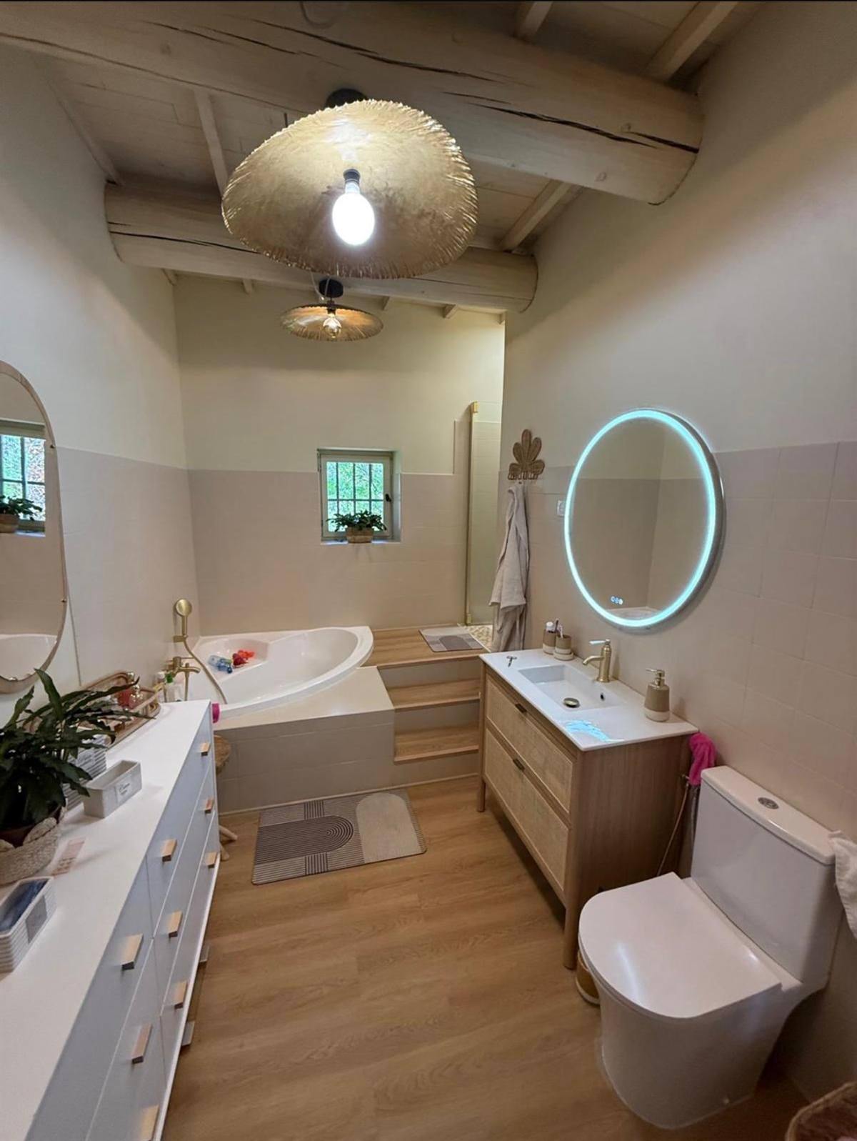 Salle de bain rénovée sous poutres apparentes : carrelage mural clair, sol aspect bois, baignoire d'angle